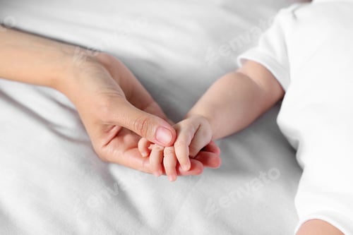 Preview: Mother holding hand of her little baby on bed, closeup