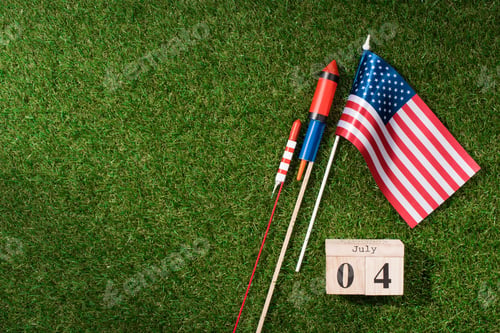 Preview: flat lay with american flagpole, wooden calendar with 4th july date and fireworks on green grass,