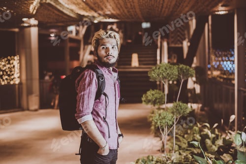 Preview: Handsome bearded man with backpack standing on the street while traveling at night.