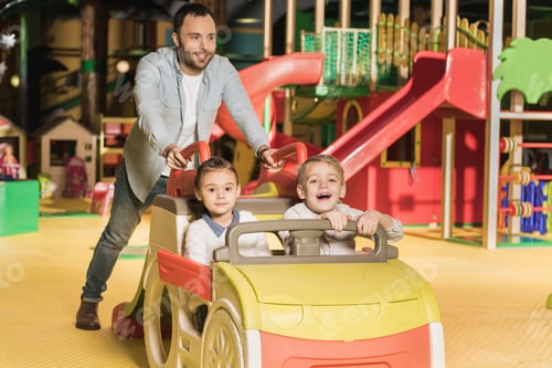 Preview: smiling father carrying adorable happy kids on toy car at entertainment center