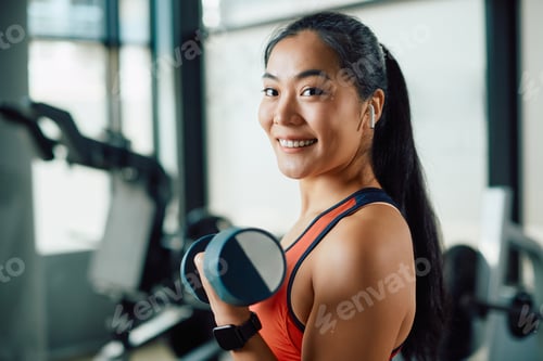 Preview: Happy Asian athletic woman exercising with hand weights in a gym and looking at camera.