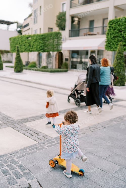 Preview: Little girl rides a scooter against the backdrop of mothers walking with strollers