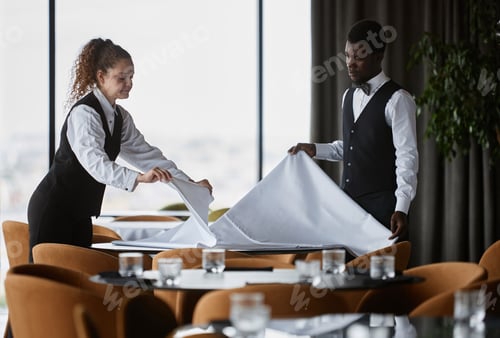 Preview: Two elegant servers setting tables with white tablecloths in restaurant