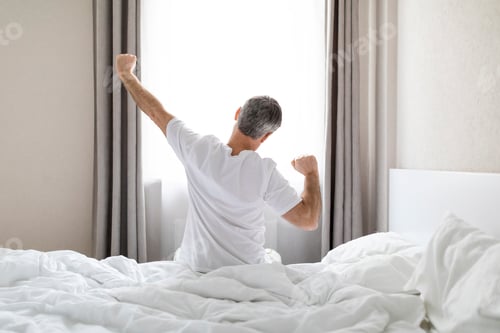 Preview: Back view of grey-haired man in pajamas stretching in bed