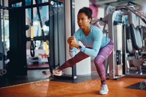 Preview: Black athletic woman practicing side lunge with suspension strap while working out in gym.