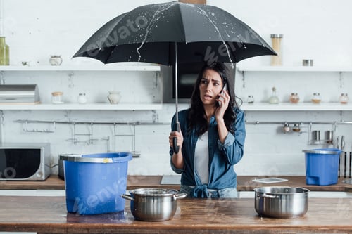 Preview: Upset woman standing under umbrella in kitchen and calling plumber