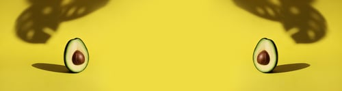 Preview: banner two cut avocado fruit stands on yellow background with shadow of monstera leaves