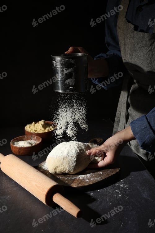 Preview: Woman sprinkling flour over dough at black table, closeup