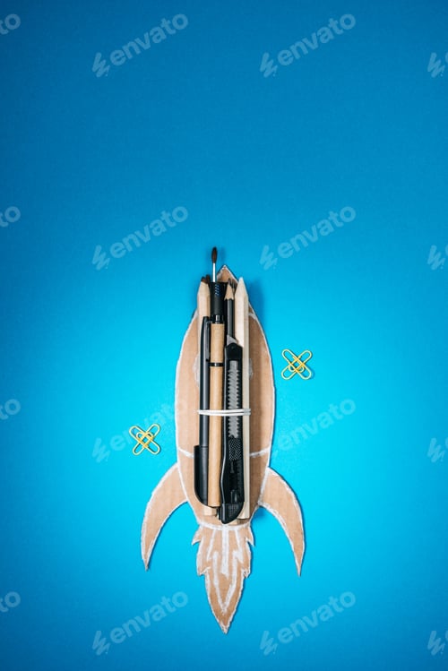 Preview: top view of handmade cardboard rocket with stationery and paper clips on blue background