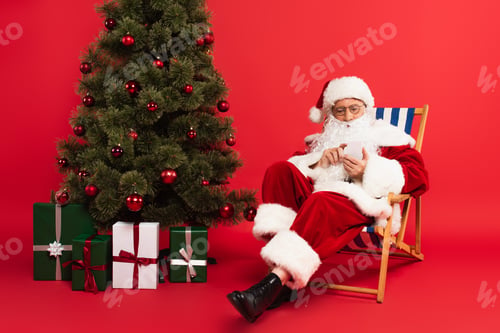 Preview: Santa claus using smartphone on deck chair near Christmas tree and gifts on red background