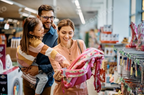 Preview: Happy little girl pointing at backpack while buying school supplies with her parents in supermarket.