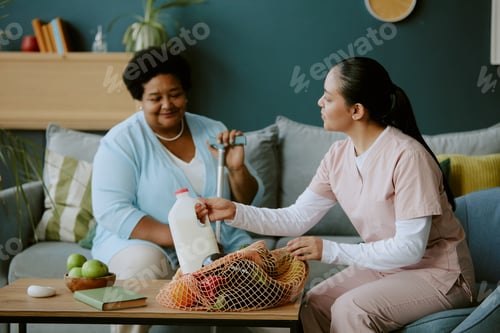 Preview: Young nurse unboxing purchase and putting them on table while senior African American female