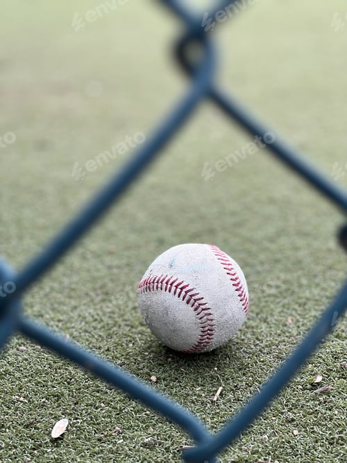 Preview: Looking though a chain link fence at a baseball with red stitching on the field. Summer day