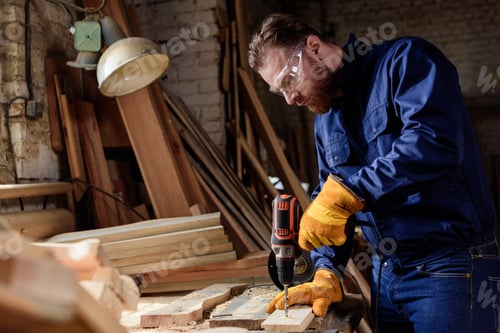 Preview: bearded carpenter in protective gloves and googles using electric drill at sawmill