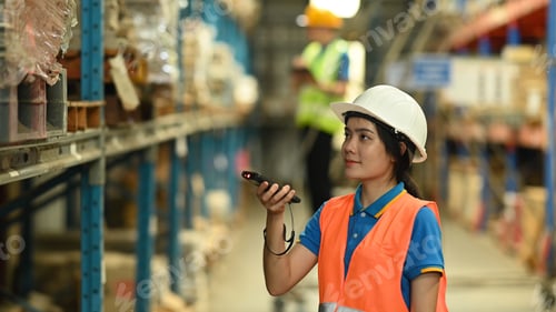 Preview: Female warehouse worker wearing safety hard hat and vest scanning barcode on box.
