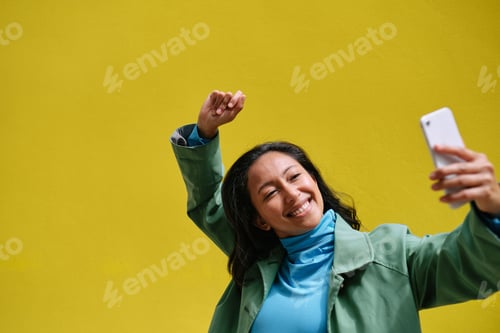 Preview: Happy young woman taking a selfie while dancing with yellow background