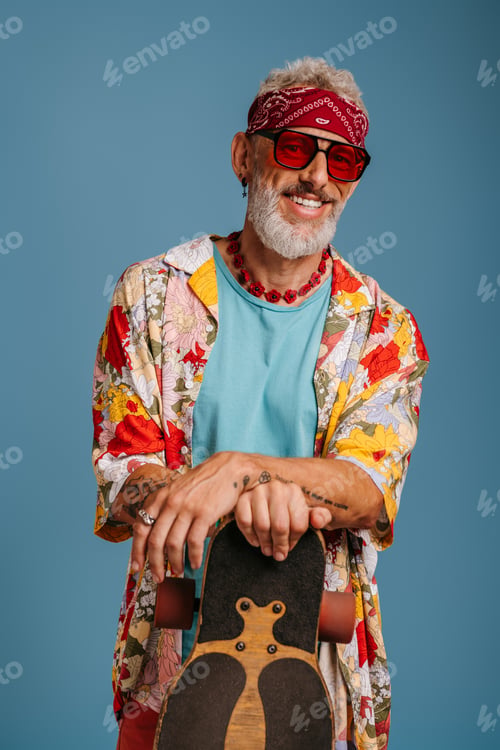 Preview: Hipster senior man in stylish funky shirt leaning on longboard and smiling against blue background