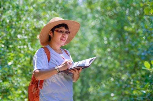 Preview: Happy Asian female tourist while taking note on nature trail in mangrove forest at natural parkland
