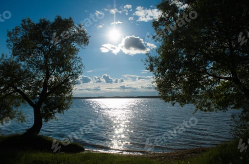 Preview: Blue cloudy sky, lake, sunbeam and trees on the sides of the frame. Outdoor daytime landscape. Glare