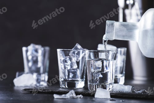 Preview: Vodka in shot glasses pouring out of the bottle on black stone background, iced strong drink
