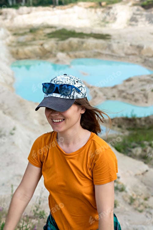 Preview: Happy Young woman in cap traveler in ginger T-shirt walking on sand of clay pit with blue water