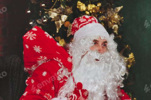 Preview: Santa Claus with bag of gifts by Christmas fireplace in decorated living room with Christmas tree