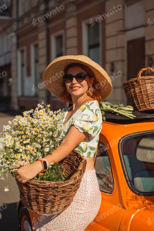 Preview: Young beautiful woman in a hat and sunglasses holding a basket of flowers, leaning on a vintage car