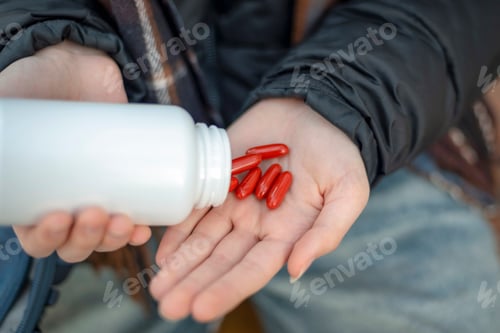 Preview: Red capsules falling from a white bottle into an open palm, captured in a close outdoor scene