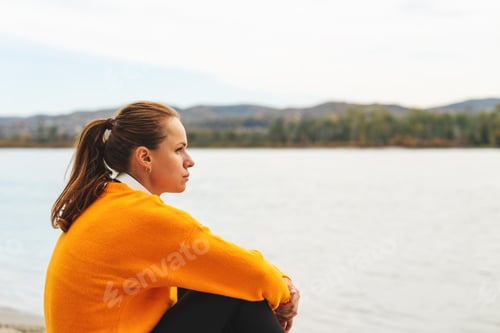 Preview: Woman sitting quietly by river during autumn season and looking into distance