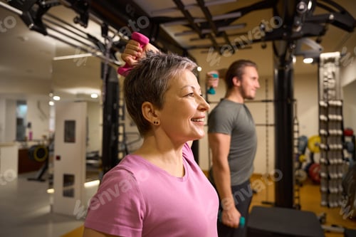 Preview: Middle aged woman and blur trainer doing exercise with dumbbells for shoulders