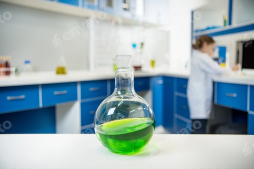 Preview: close up view of glass tube wit liquid on lab table with scientist behind