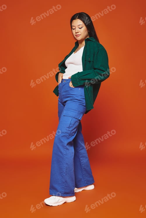 Preview: stylish young asian woman posing in vibrant outfit with hands in pockets on orange background