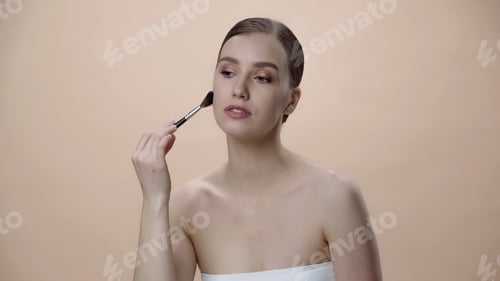 Preview: woman with bare shoulders applying face powder while holding cosmetic brush isolated on beige