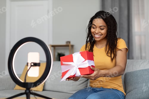 Preview: Happy black woman holding gift box and shooting video for blog, looking at cellphone set on ring