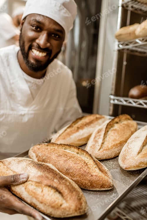 Preview: smiling handsome african american baker with tray of fresh loaves of bread on baking manufacture
