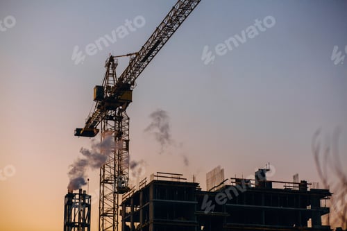 Crane and building construction site with pipe with smoke on background of sunset sky