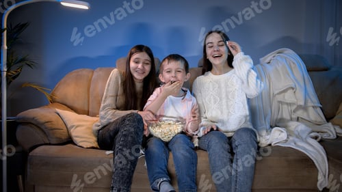 Preview: Smiling little boy with two teenage girls watching late night TV show and eating popcorn from big