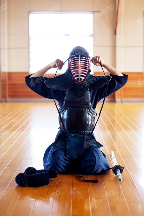 Preview: Female Japanese Kendo fighter kneeling on wooden floor, fastening Kendo mask.