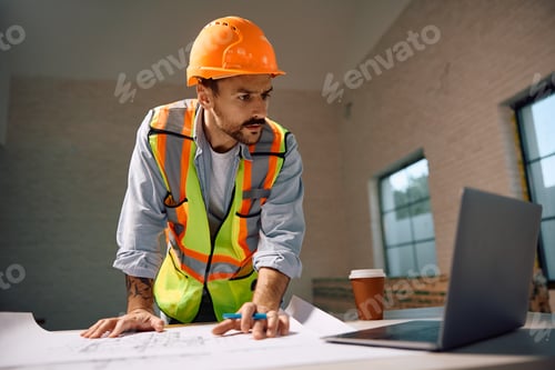 Preview: Construction worker reading housing plans while using laptop during home renovating process.