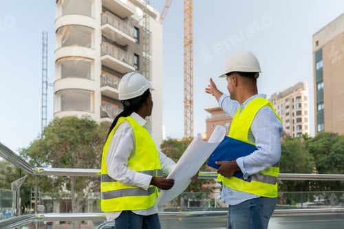 Preview: Two Diverse architects in safety vests discussing building project at urban construction site