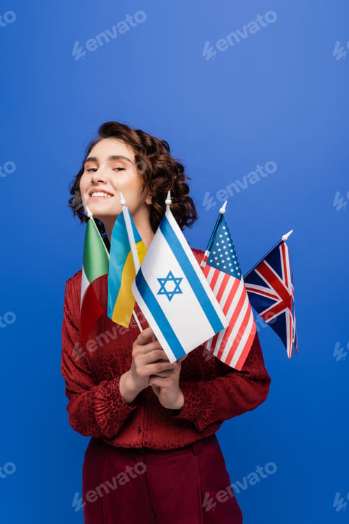 Preview: young inspired woman looking at camera while holding flags of different countries isolated on blue