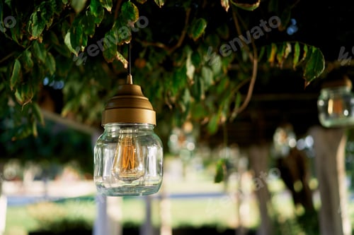 Preview: Jar lamp hangs from the ceiling of a pergola entwined with green branches in the garden