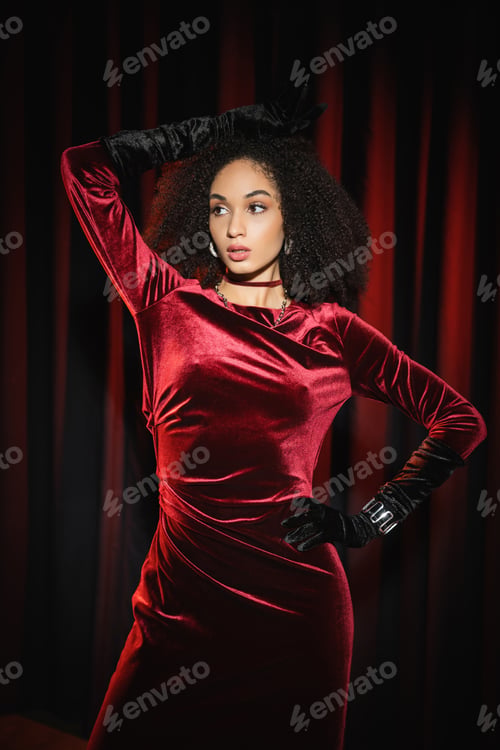 Preview: Stylish african american woman in gloves and dress looking away while posing near red drapery