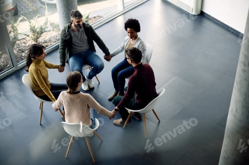 Preview: Above view of group therapy attenders holding hands while sitting in a circle.