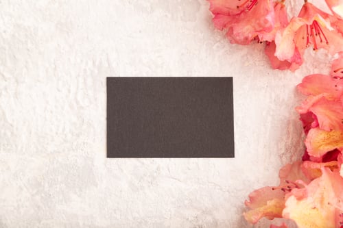 Preview: Black business card with pink azalea flowers on gray concrete background. top view, copy space.