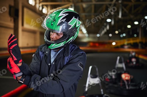 Preview: african american motorsports driver in helmet wearing gloves and standing near circuit, go-kart