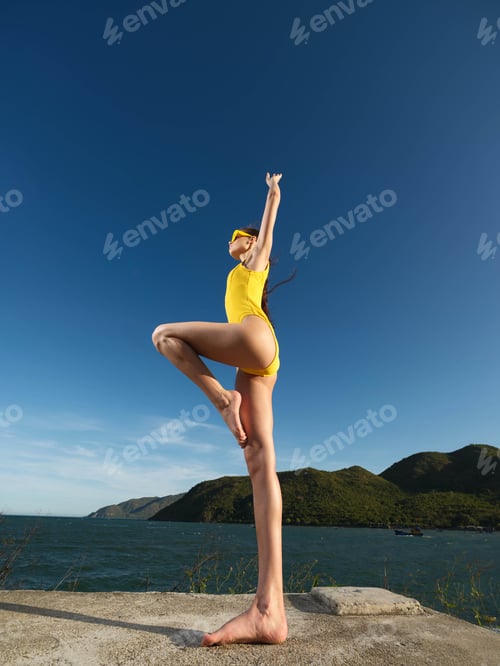 Preview: Young woman, yellow swimsuit, handstand on rocky cliff edge, ocean backdrop, blue sky, athletic pose