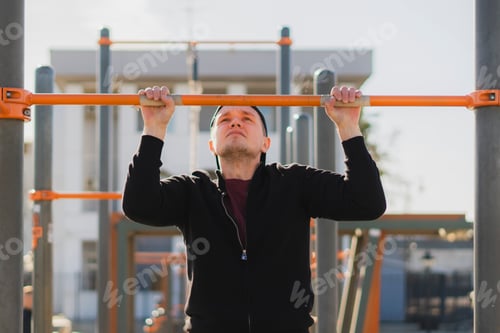 Preview: handsome healthy athletic guy doing callsthenics pull-ups outdoor on a sports ground