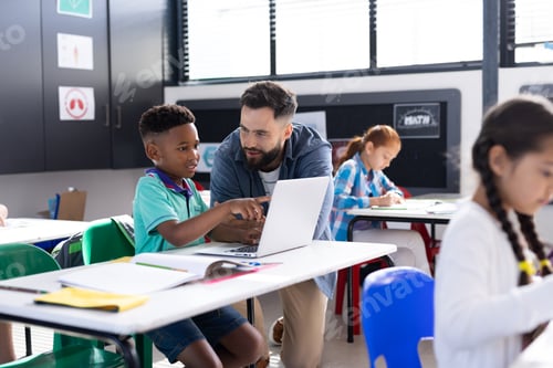 Preview: Happy male teacher and schoolboy using laptop at his desk in diverse elementary class, copy space