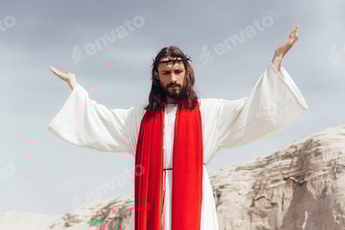 Preview: low angle view of Jesus in robe, red sash and crown of thorns standing with raised hands in desert